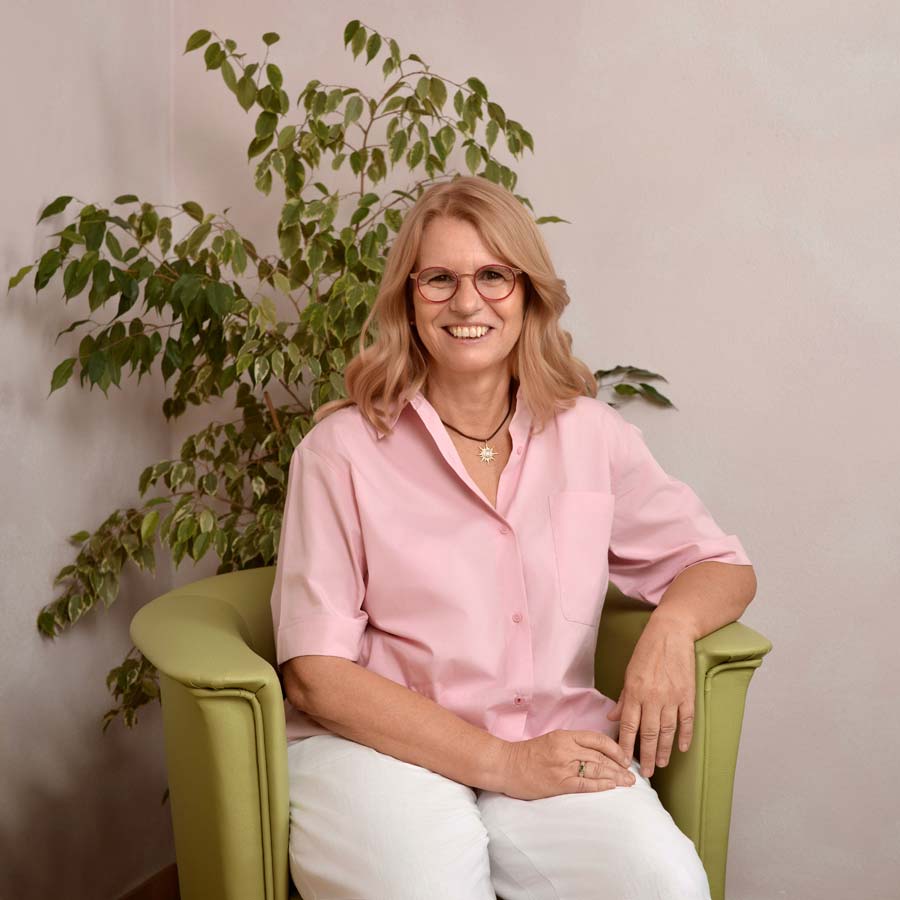 Doris Borowski sitzt lächelnd in einem hellgrünen Sessel, in rosa Bluse und weisser Hose, hinter ihr eine grosse Zimmerpflanze.
